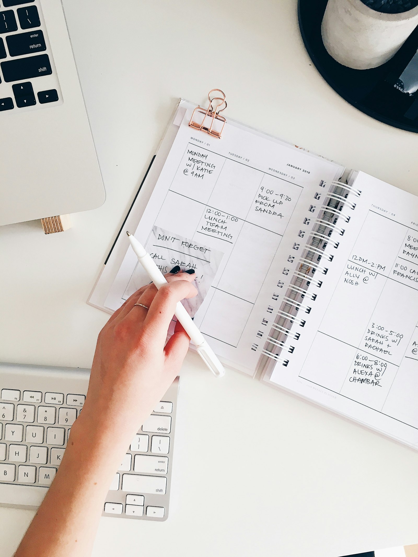 Hand writing in a weekly planner on a white desk beside a laptop keyboard
